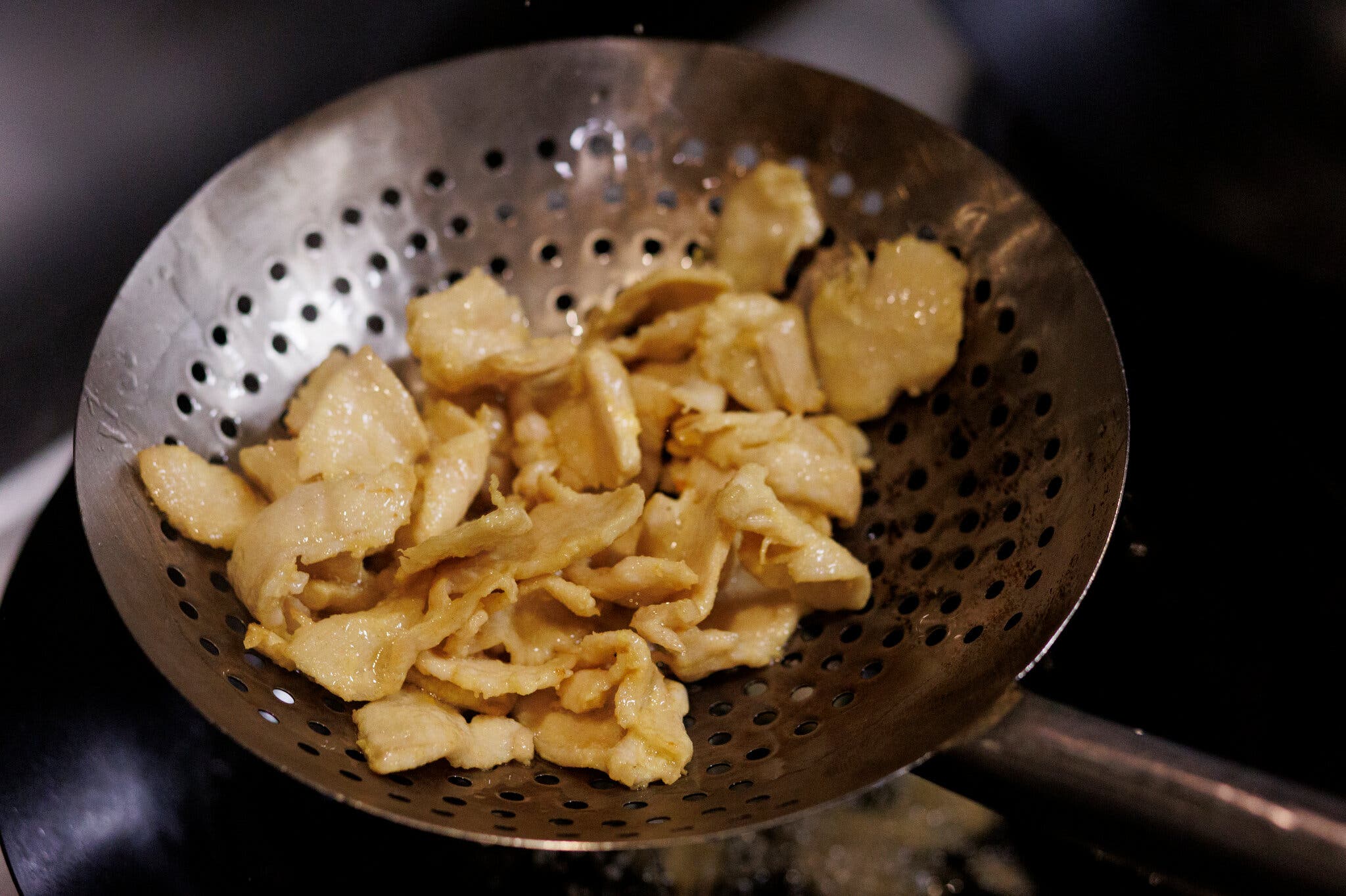 A large strainers holds shiny slices of chicken breast.