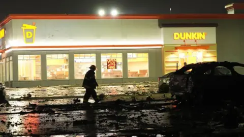 Getty Images A fireman walks down a street littered with debris outside a branch of Dunkin' Donuts. A burnt out car can be seen ahead of him