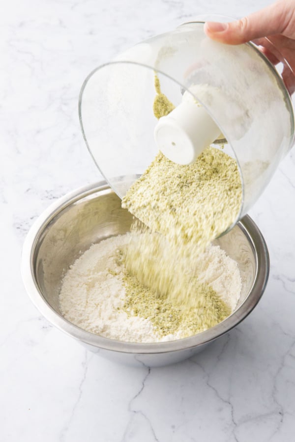 Pouring finely ground pistachios into a mixing bowl with flour and dry ingredients.