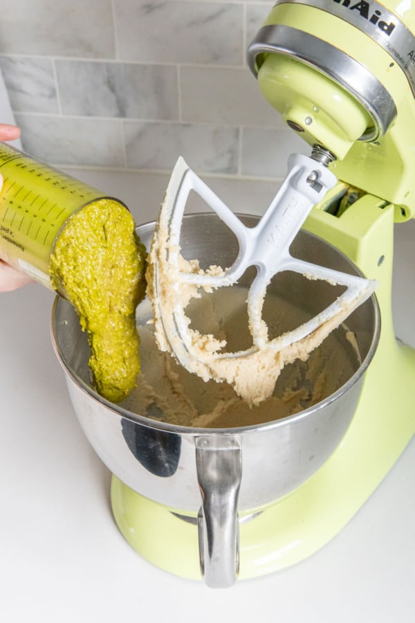 Adding bright green pistachio butter to a mixer bowl with creamed butter and sugar.