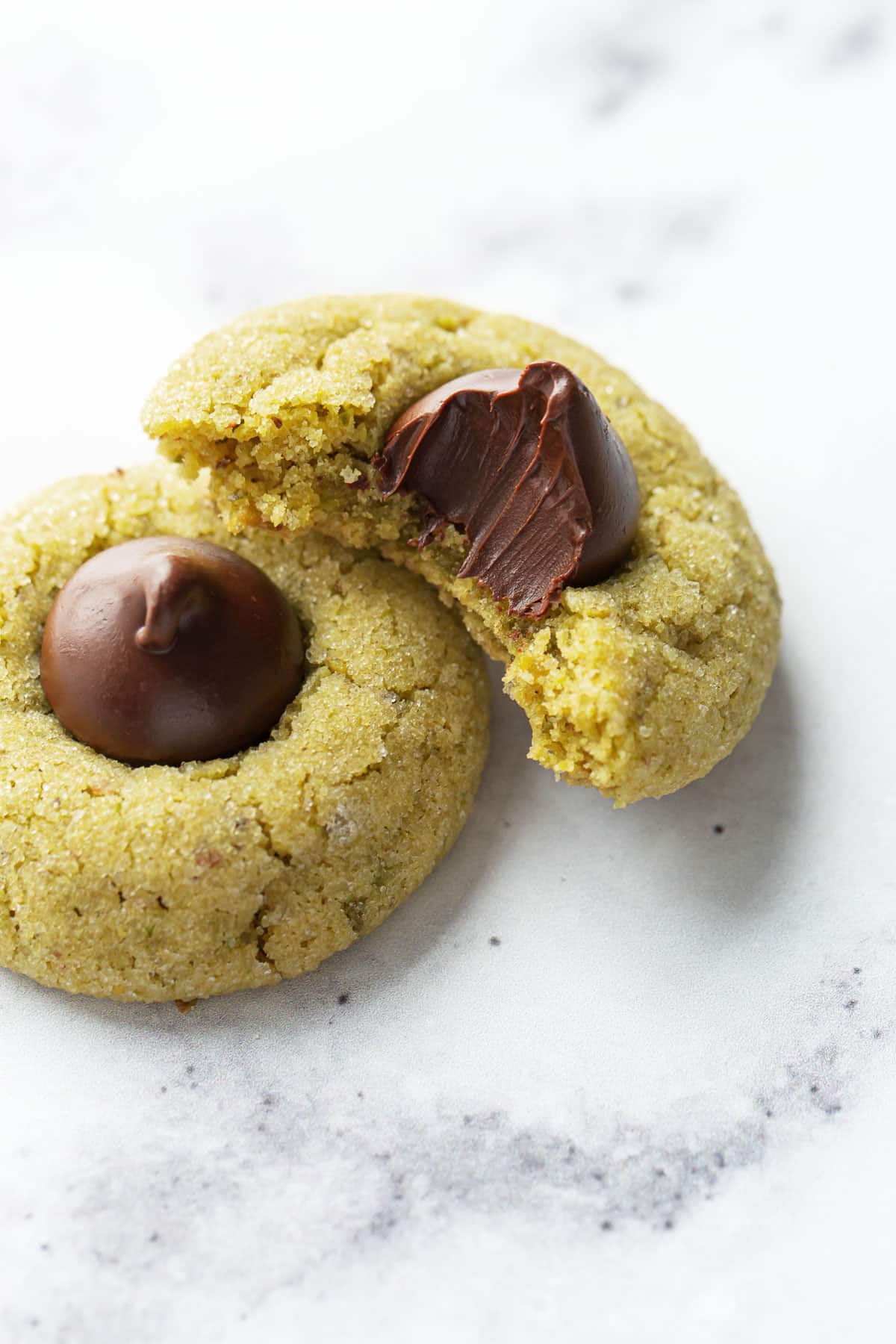 Closeup, two Pistachio Butter Blossom Cookies on a marble background, one cookie with a bite taken out to show the cookie's texture.