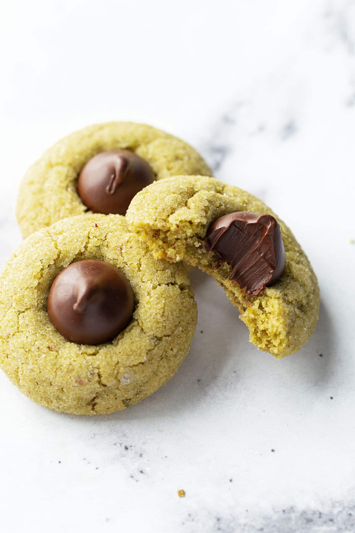 Pile of three Pistachio Butter Blossom Cookies on a marble background, one cookie with a bite taken out to show the cookie's texture.