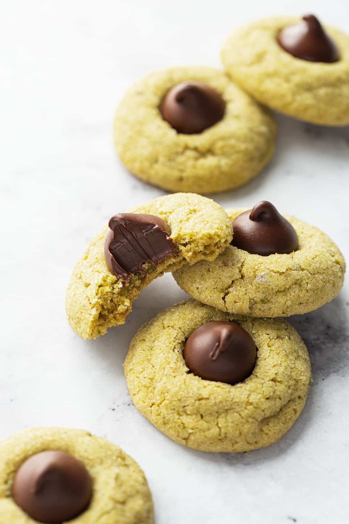 Scattered Pistachio Butter Blossom Cookies on a white background, one cookie with a bite out of it to show the dark chocolate and soft texture of the cookie.