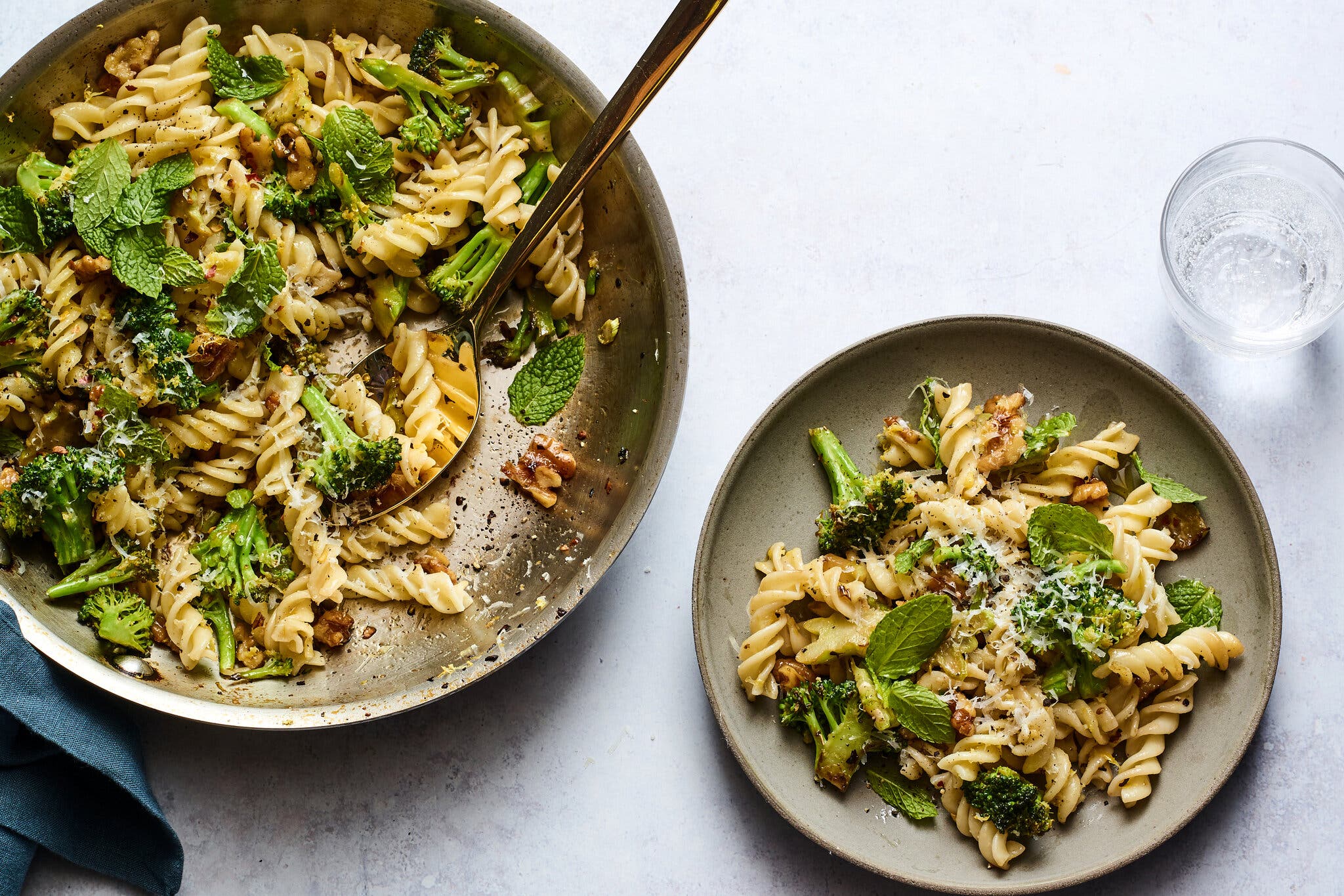 A skillet filled with fusilli pasta, herbs and broccoli sits next to a single serving.