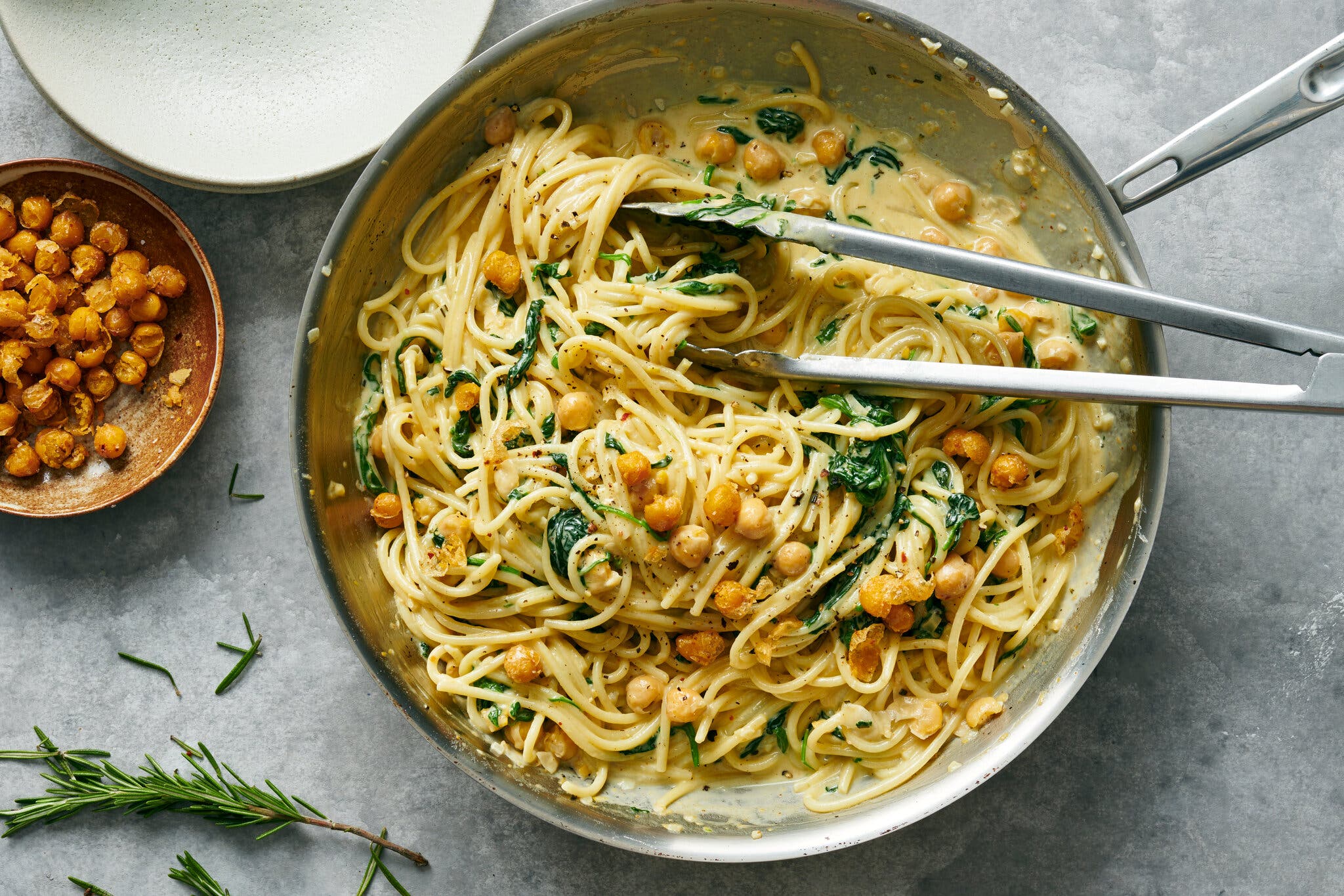 An overhead image of saucy spaghetti, chickpeas and greens in a skillet set underneath some tongs. A small bowl of crisped chickpeas sits off to the side.