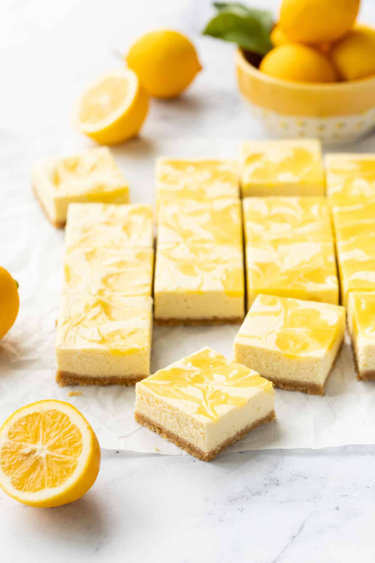 Cut squares of Meyer Lemon Cheesecake Bars with a swirl of lemon curd on top, on a piece of parchment with a few cut and whole Meyer lemons and a bowl of lemons in the background.