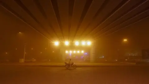 Getty Images A lone man rides his motorbike through an orange haze with rows of bright street lights shining overheard in Najaf, Iraq on 14 April 2025. He rides underneath a bridge with long rows of planks stretching out towards the foreground.