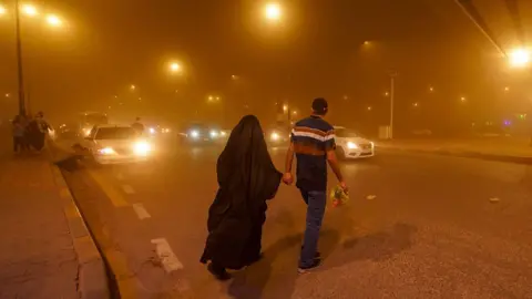 Getty Images During a sandstorm in Najaf, Iraq on 14 April 2024, a man dressed in a black shirt with red and white stripes walks hand in hand with a woman dressed in a black burka. They are crossing a busy road with cars as thick, orange cloud blankets the air and lights from street lamps shimmer around them.