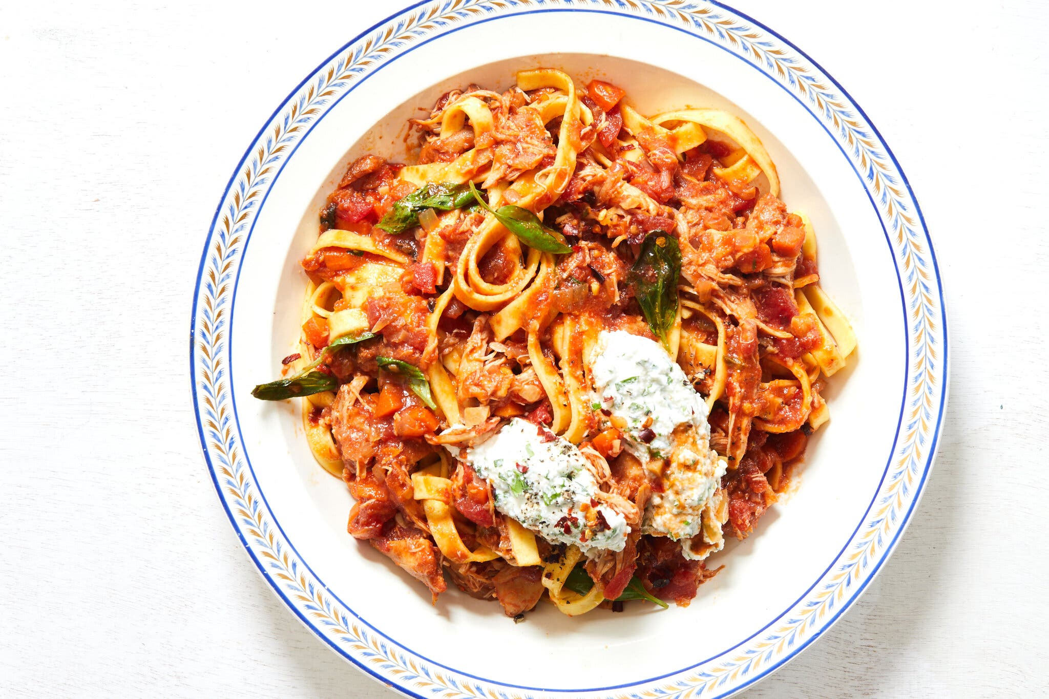 Thick noodles coated in a tomato sauce and topped with a few herbs and a creamy herbed ricotta sit in a crisp white bowl with blue and orange trim.
