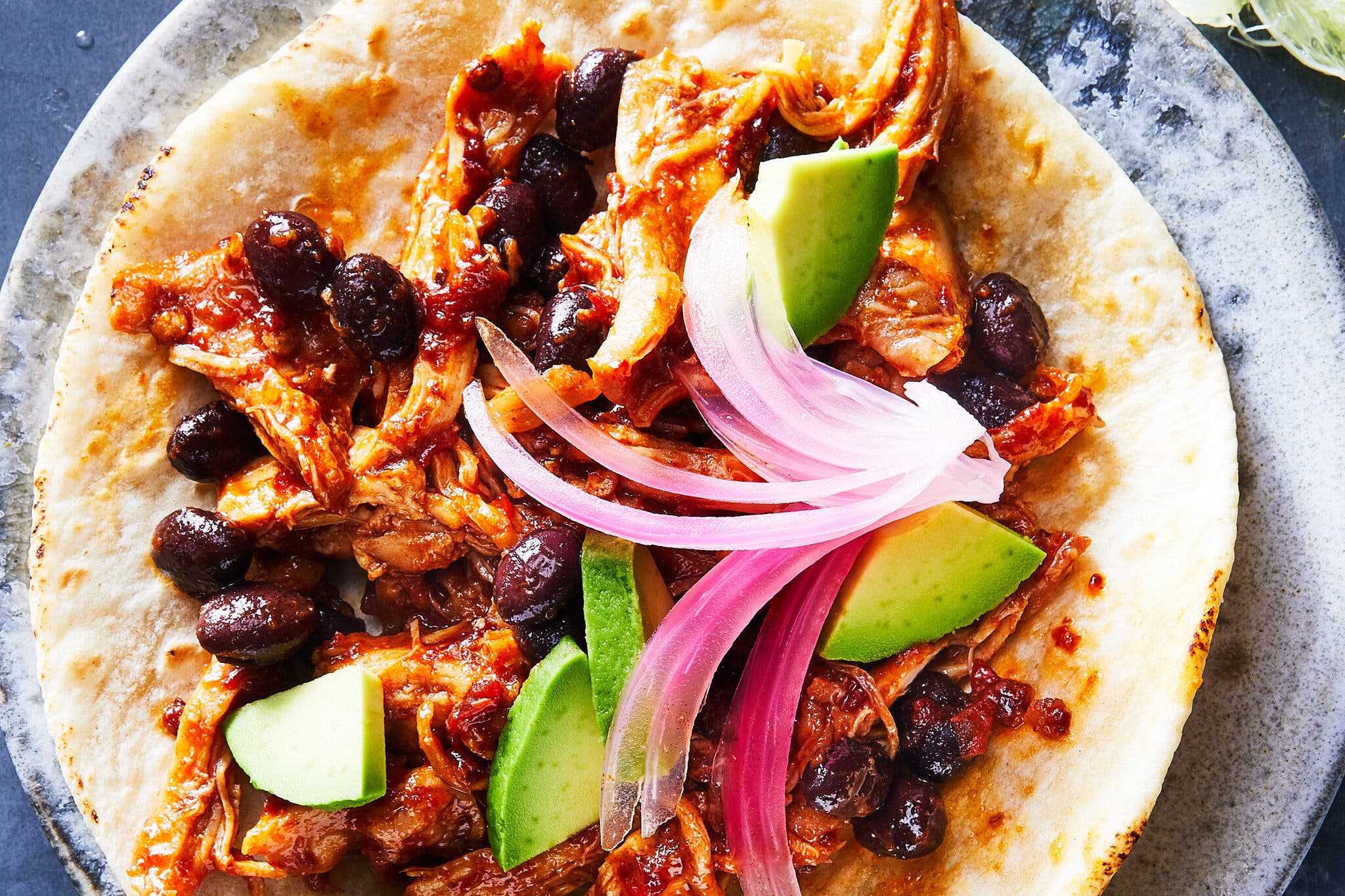 An overhead image of an open-faced flour tortilla filled with shredded chicken in red sauce, black beans, pickled red onion slices and avocado cubes. 