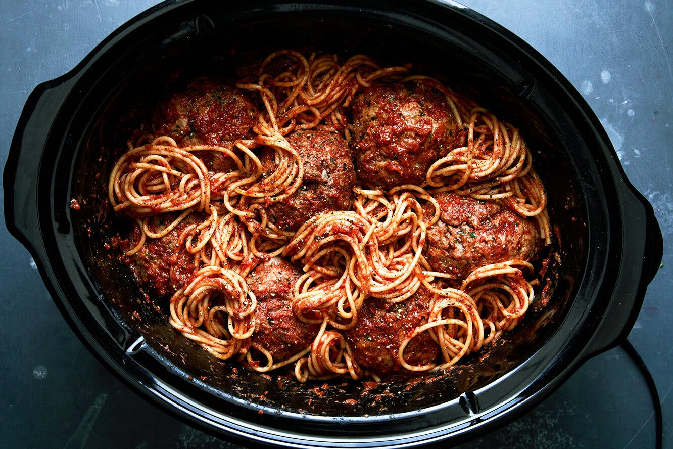 An overhead image of a black slow cooker filled with spaghetti and meatballs.
