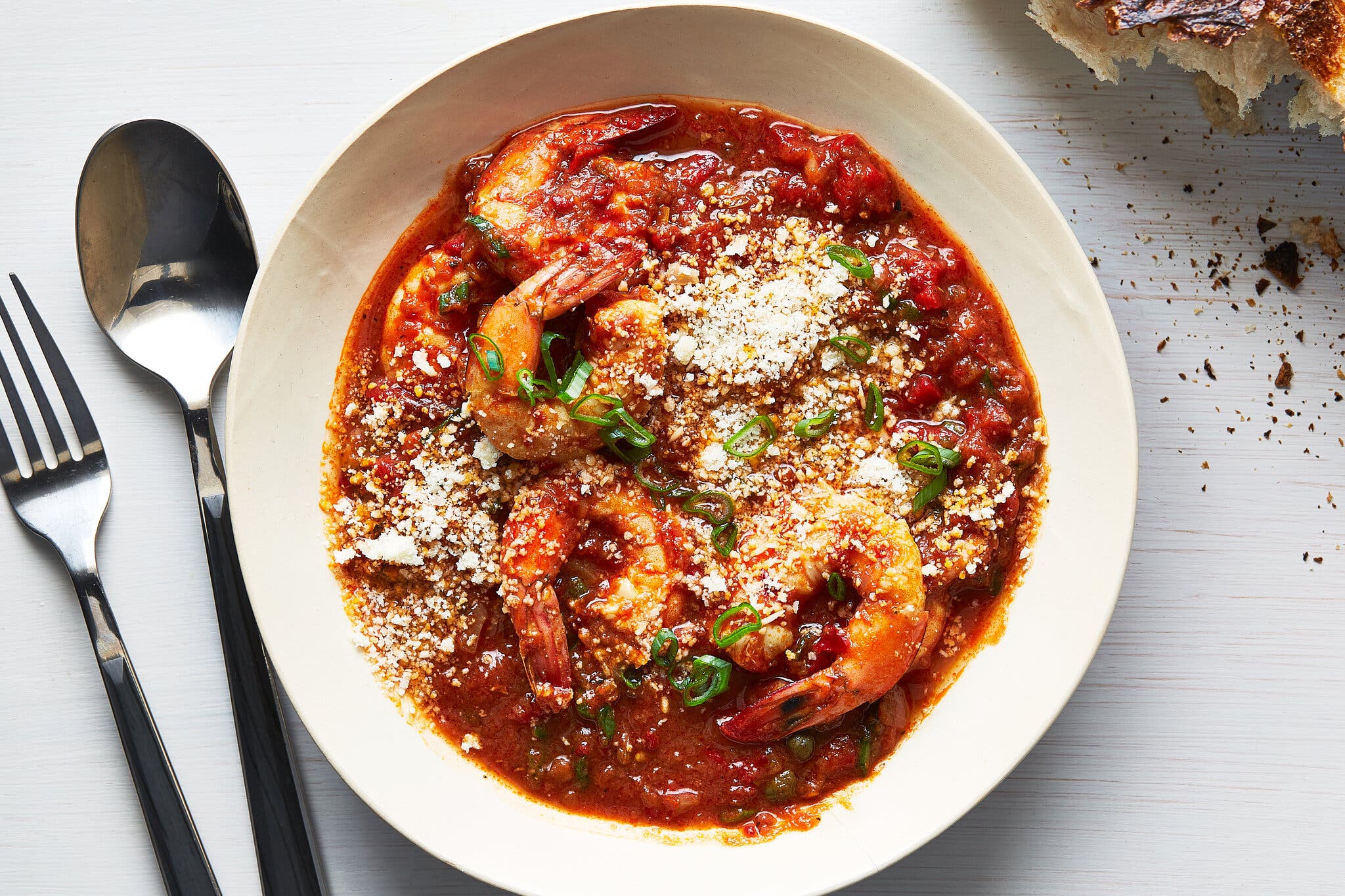 An overhead image of a white bowl filled with brick red sauce dotted with shrimp and scattered with scallions and grated Parmesan. A silver fork and spoon are to the left, and a torn piece of bread peeks into the upper right corner of the image.