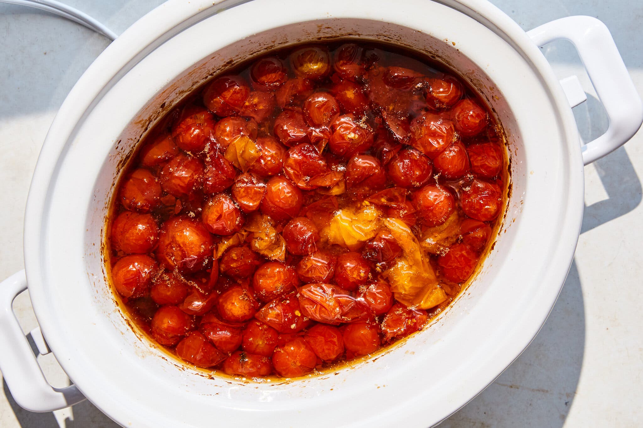 An overhead image of a white slow cooker filled with bright red cherry tomato compote on a marble countertop.