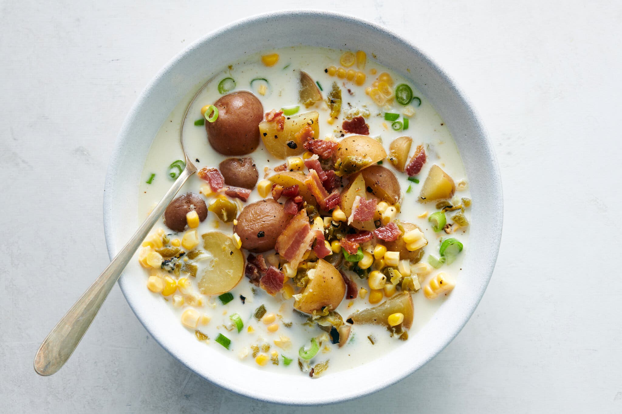 An overhead image of a white bowl against a white background, filled with a creamy soup of corn kernels, chunks of red potatoes, chopped scallions and bacon bits. 