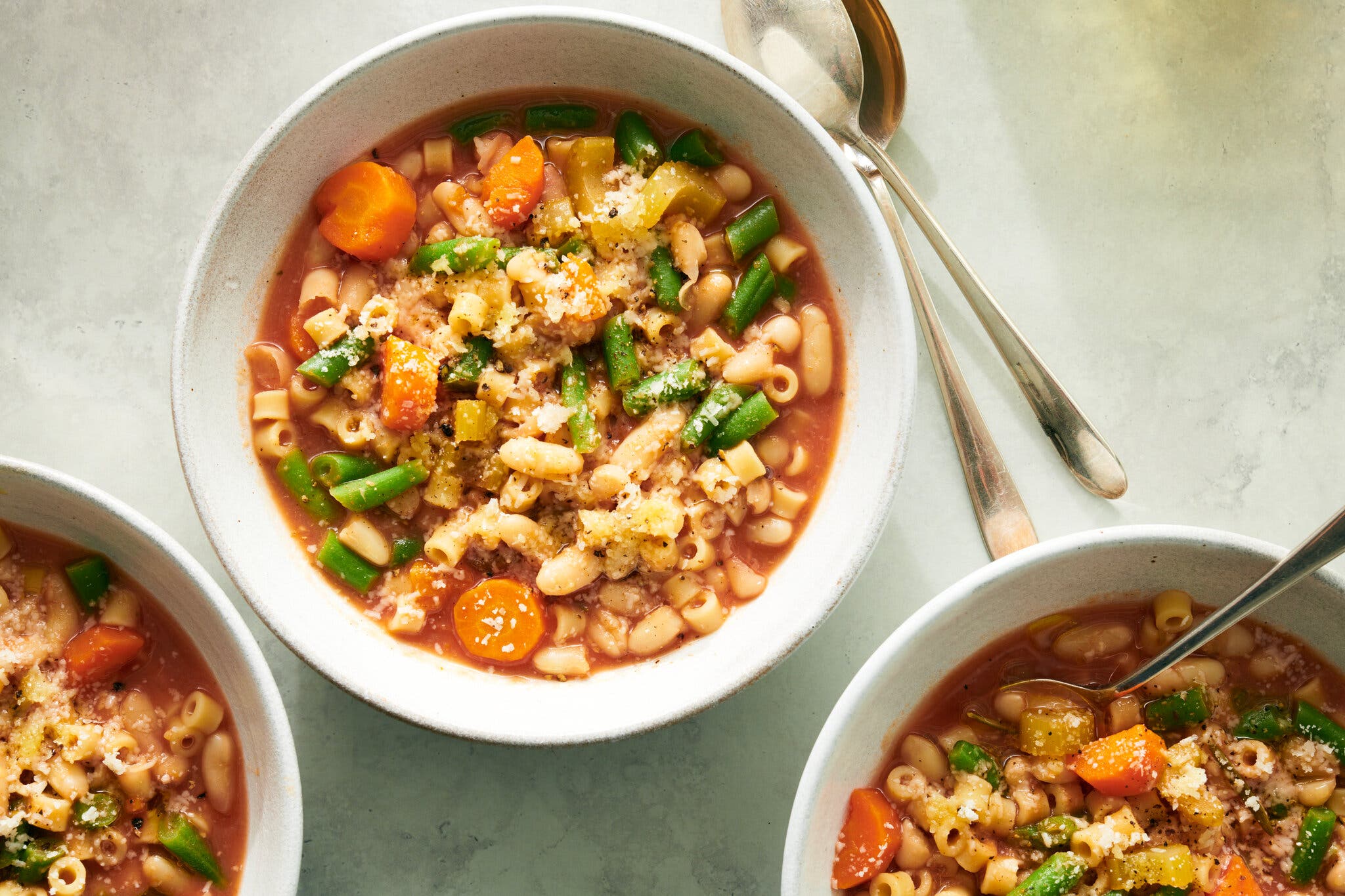 An overhead image of three white bowls filled with small pasta, white beans, chopped green beans and carrot rounds in a brick-red broth, sprinkled with grated Parmesan cheese.