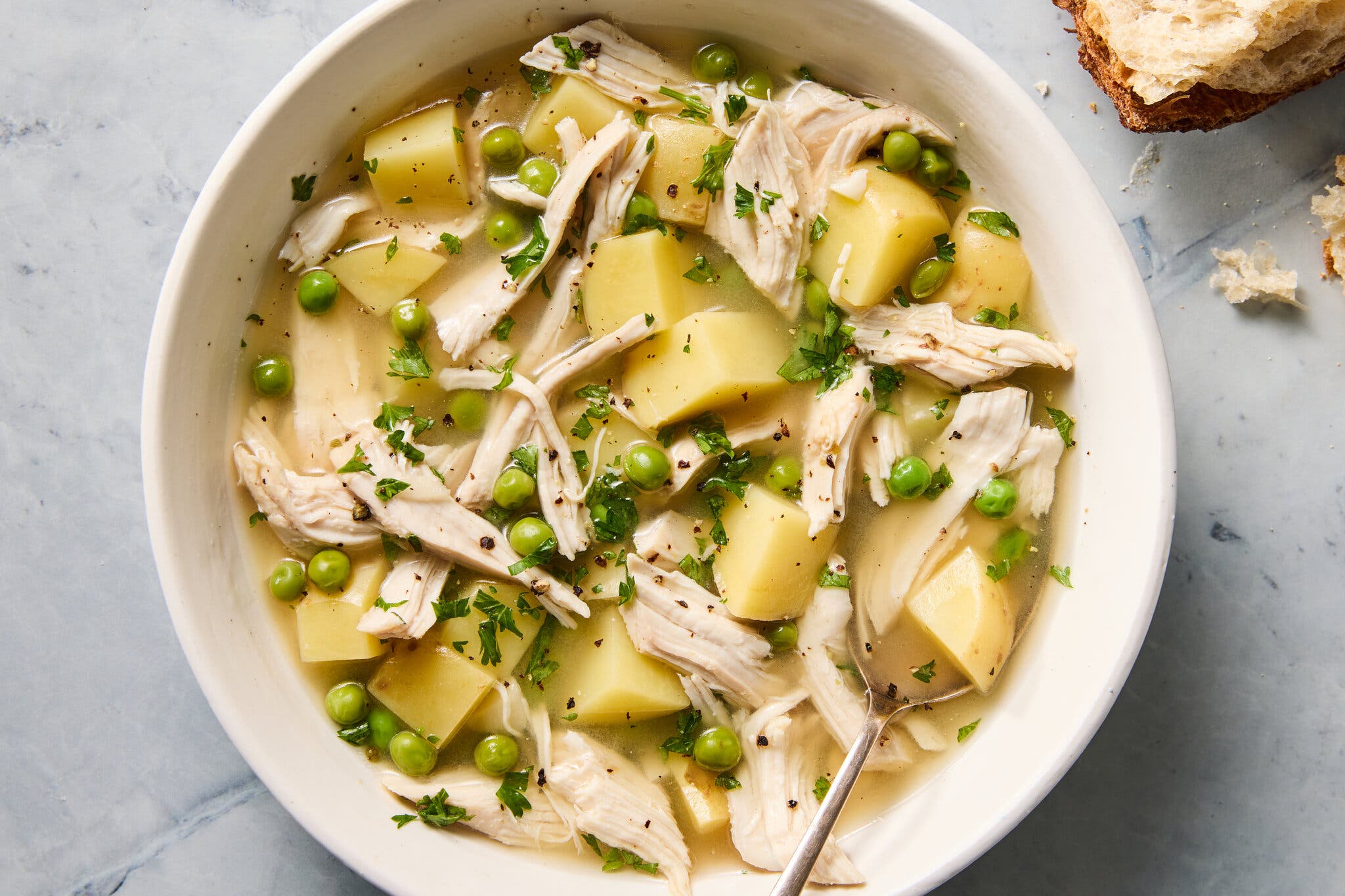 An overhead shot of a white bowl filled with a clear broth, large cubes of potatoes, shredded chicken and green peas, garnished with parsley.