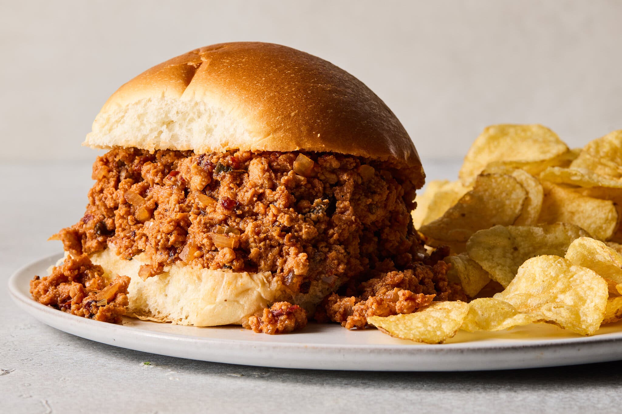 A sloppy joe sandwich, filled with a large amount of ground meat and sauce, sits on a plate next to a pile of potato chips.