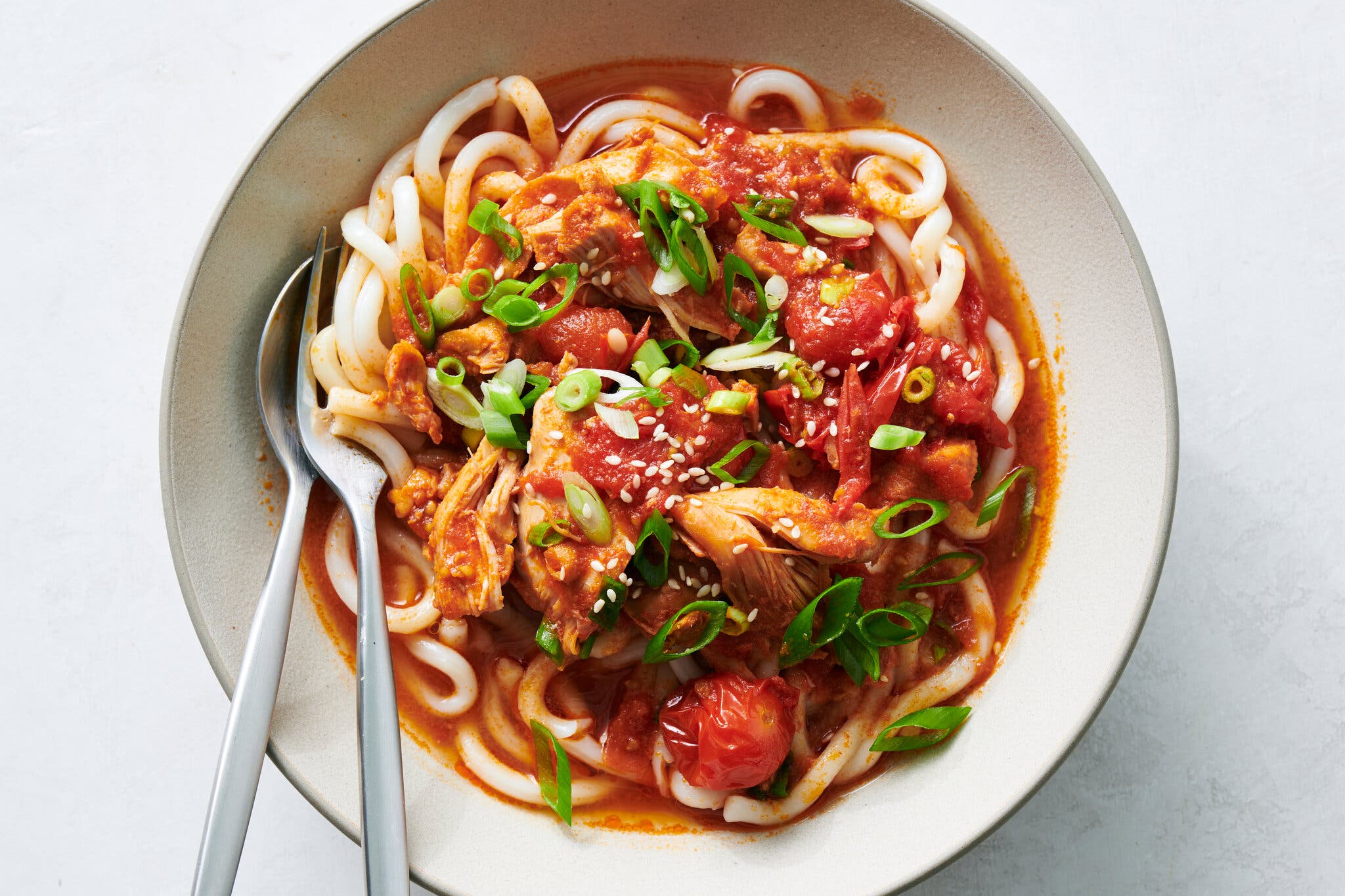 An overhead shot of a bowl of thick udon noodles with chicken, tomatoes and green onions in a red sauce, with a fork and spoon.