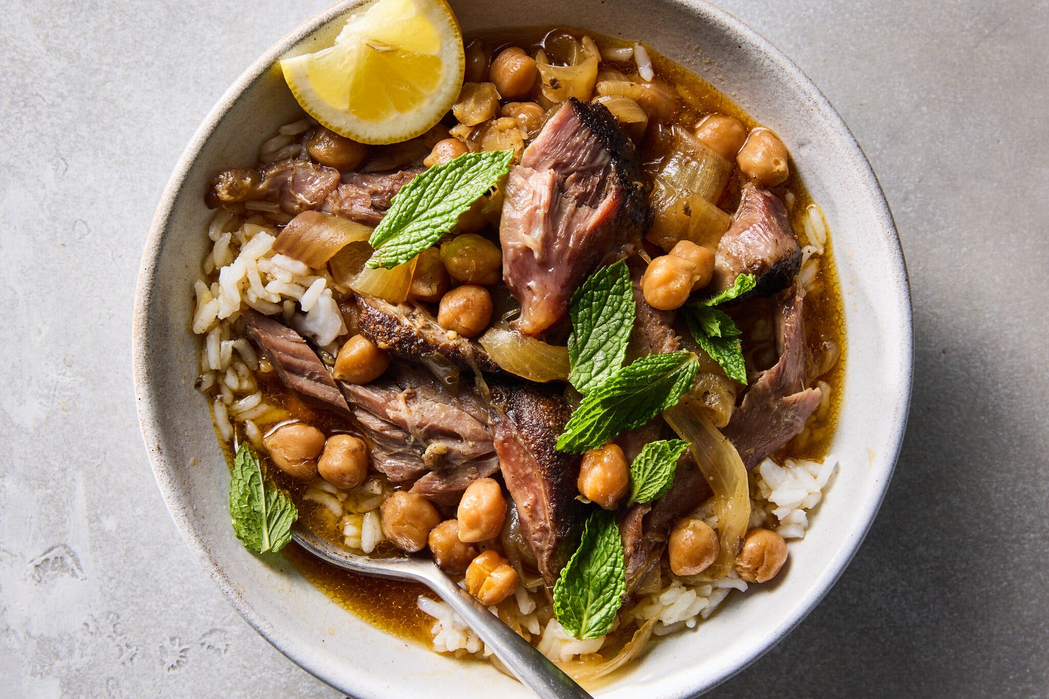 An overhead shot of a bowl of lamb stew, rice and chickpeas, garnished with fresh mint and a lemon wedge.