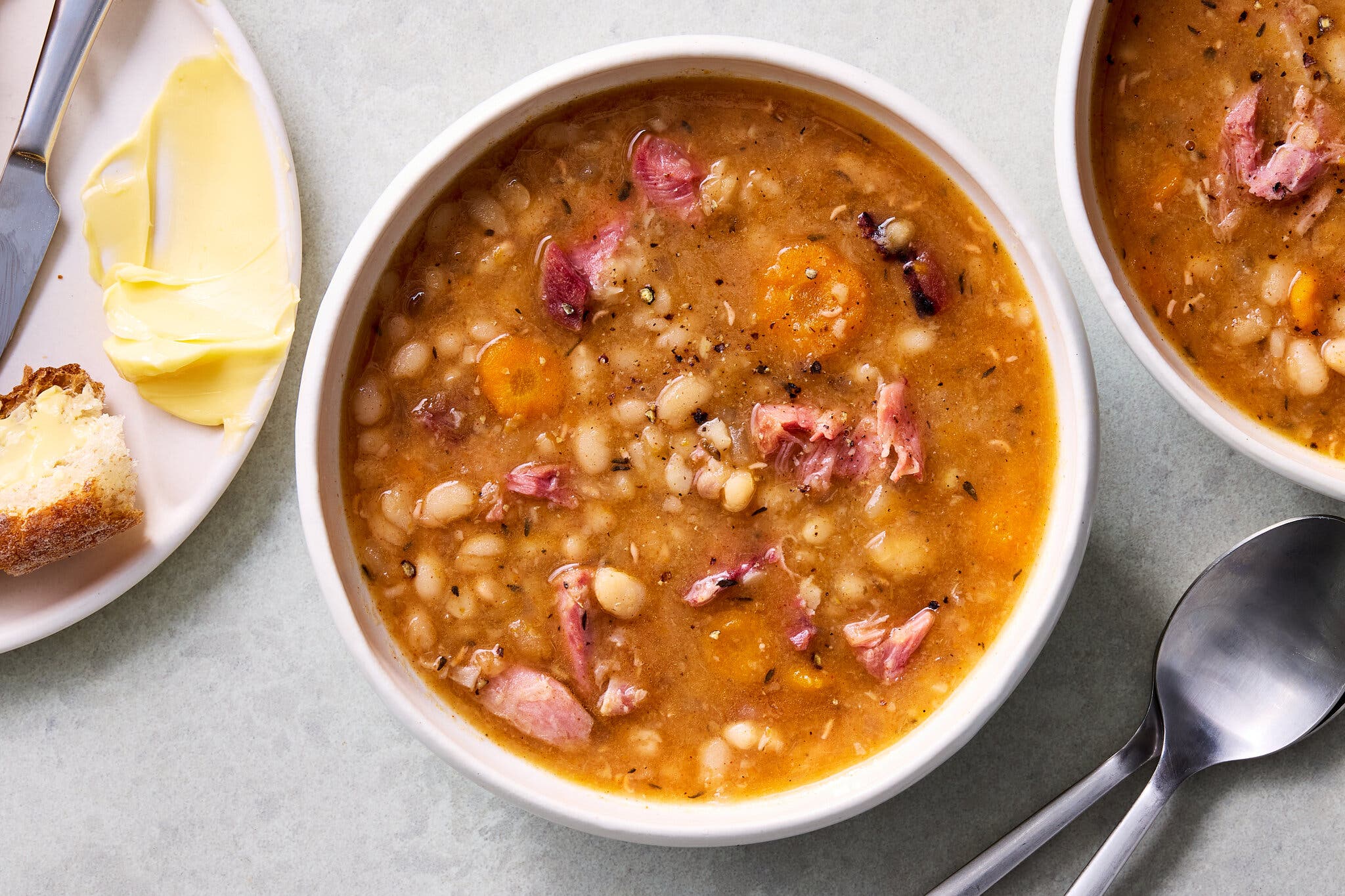 An overhead shot of a bowl of ham and bean soup with sliced carrots in a savory broth, served with a side of buttered bread.