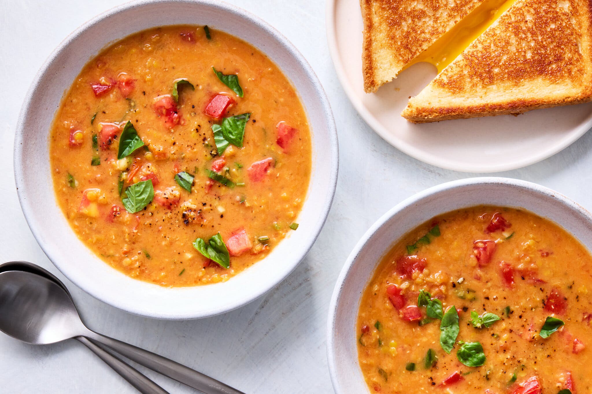 An overhead shot of two bowls of tomato and corn soup with basil, alongside a plate of grilled cheese sandwiches.