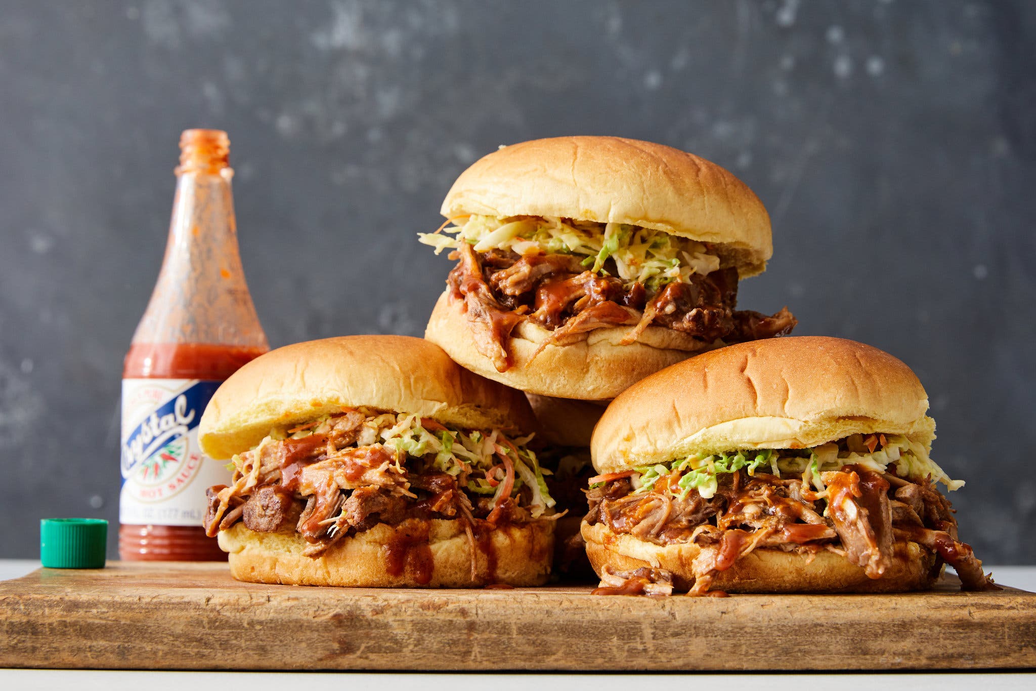 A side image of a stack of three hamburger buns filled with pulled pork and shredded lettuce. An uncapped bottle of Crystal hot sauce is to the left. 