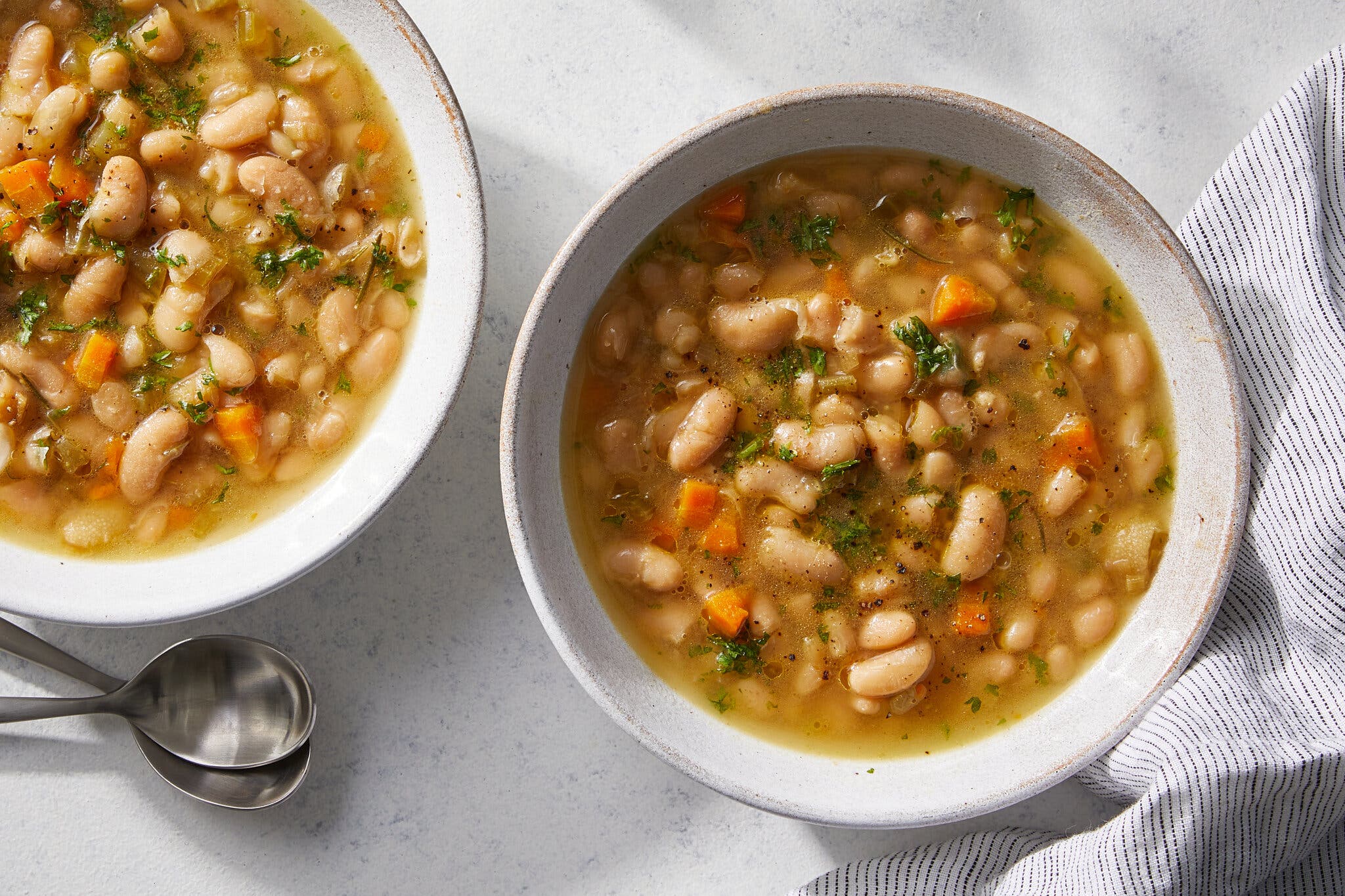 An overhead image of two white bowls filled with brothy white beans dotted with green herbs and diced carrots.
