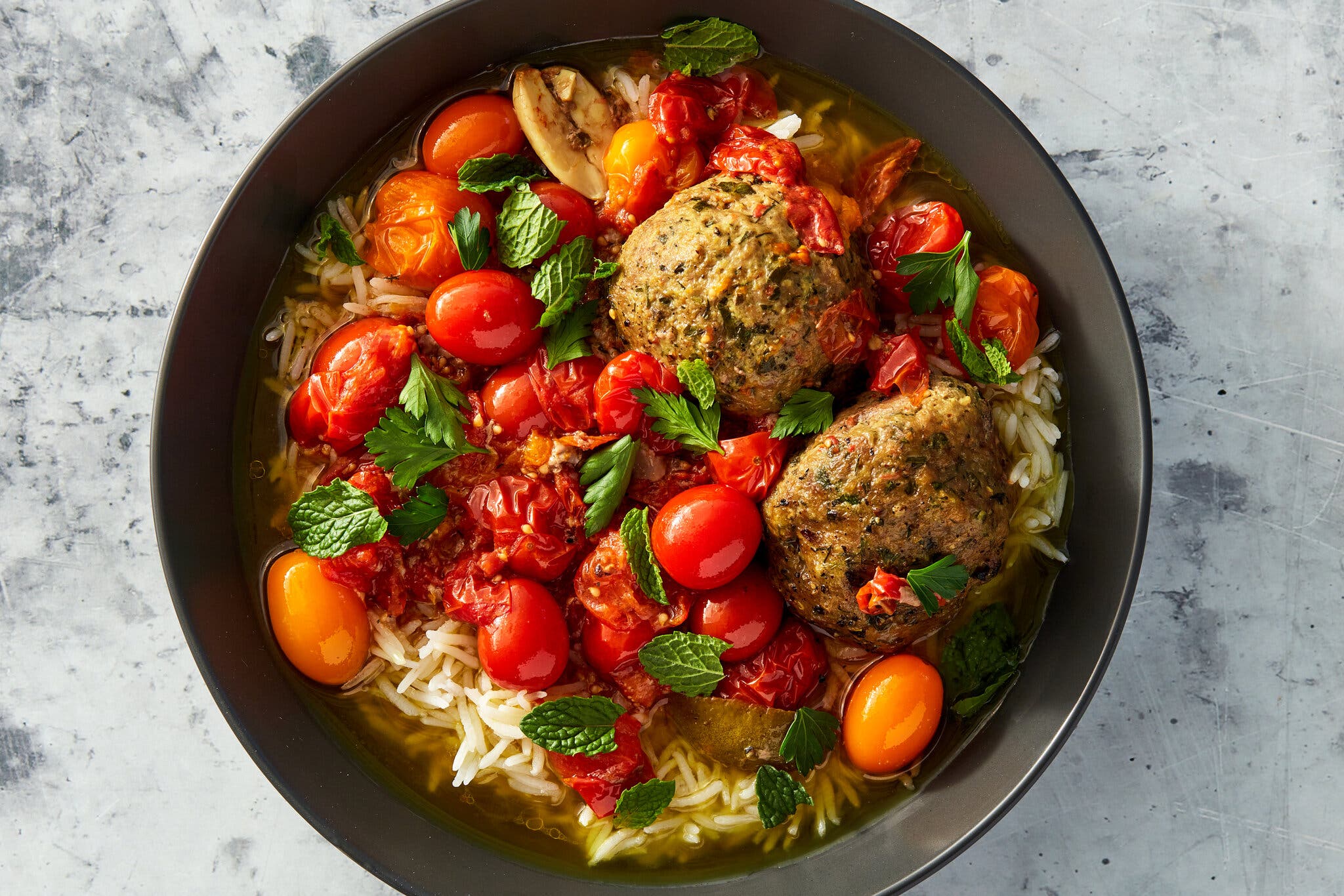 Two kofte sit atop a dark bowl strewn with herbs and cooked-down tomatoes. At the bottom of the bowl is orzo in broth.