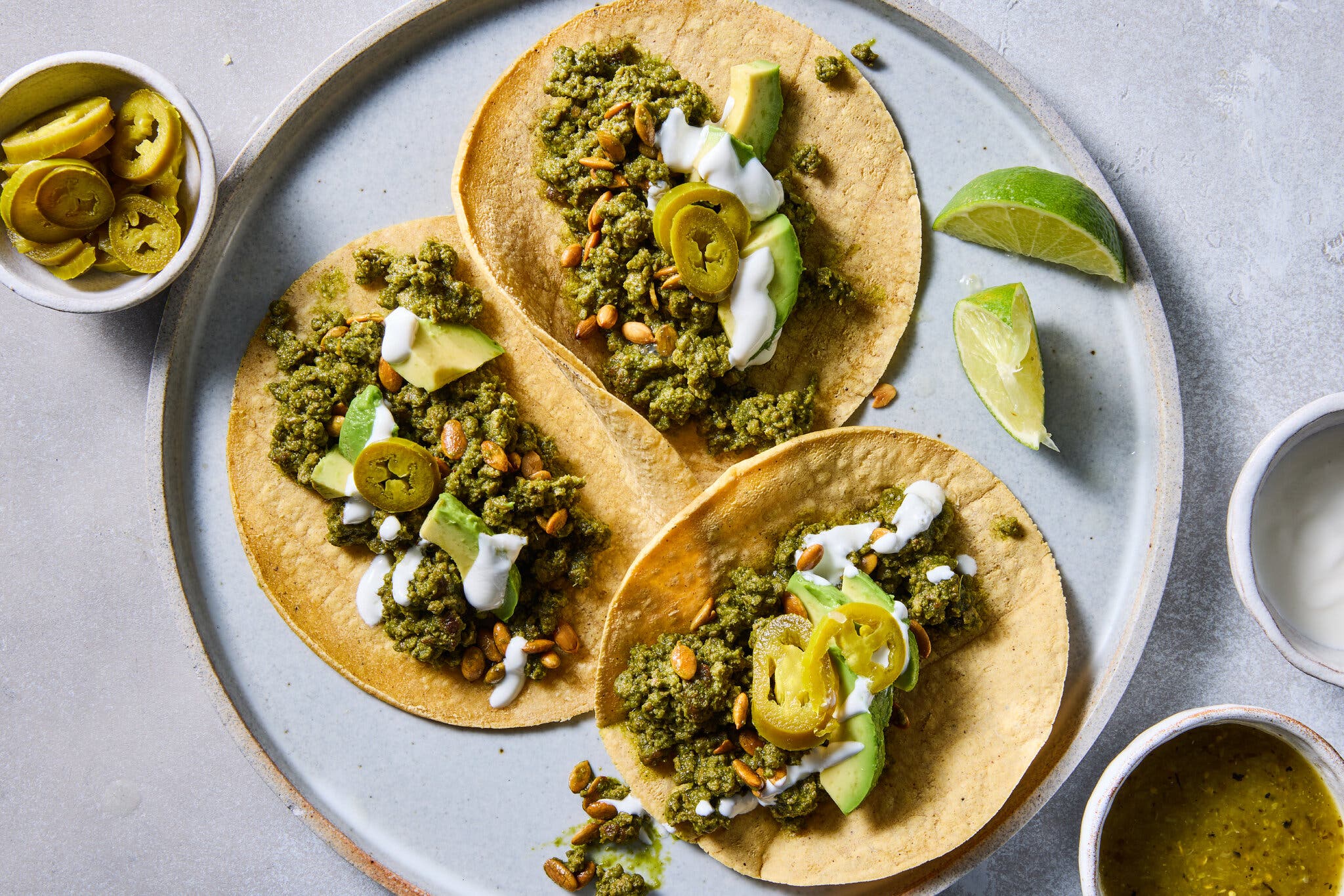 An overhead view of a light gray plate holding three tacos filled with a dark green ground beef mixture, pepitas, avocado slices and sour cream; a bowl of jalapeños and lime wedges sit nearby.