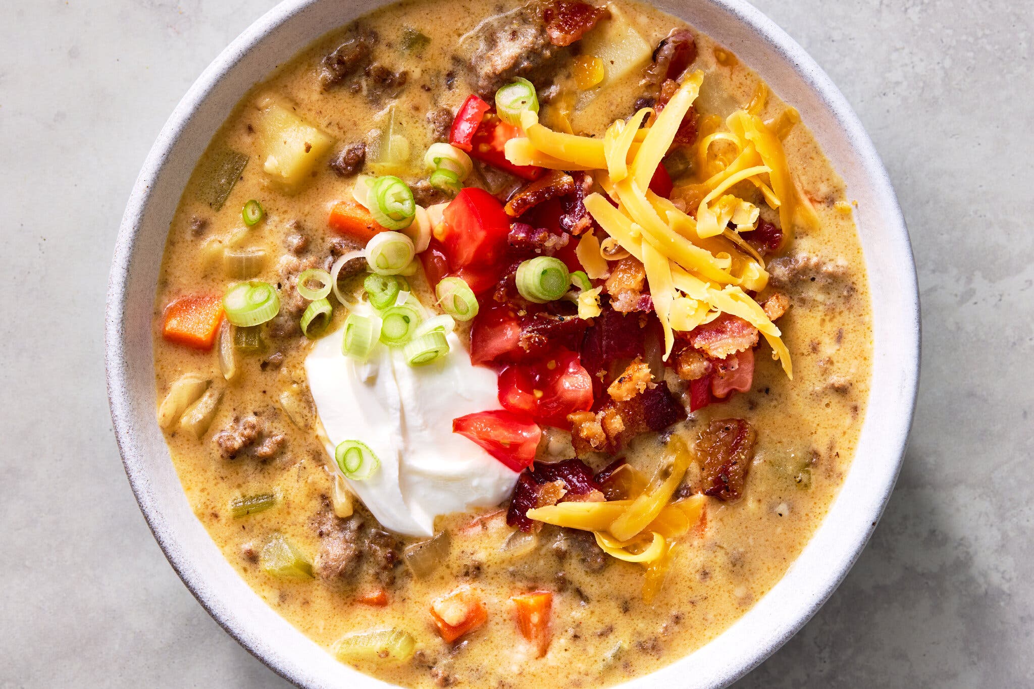 An overhead close-up of a bowl of rich, brown-and-gold colored cheeseburger soup, garnished with a dollop of sour cream, bacon crumbles, cheese shreds and green onions.