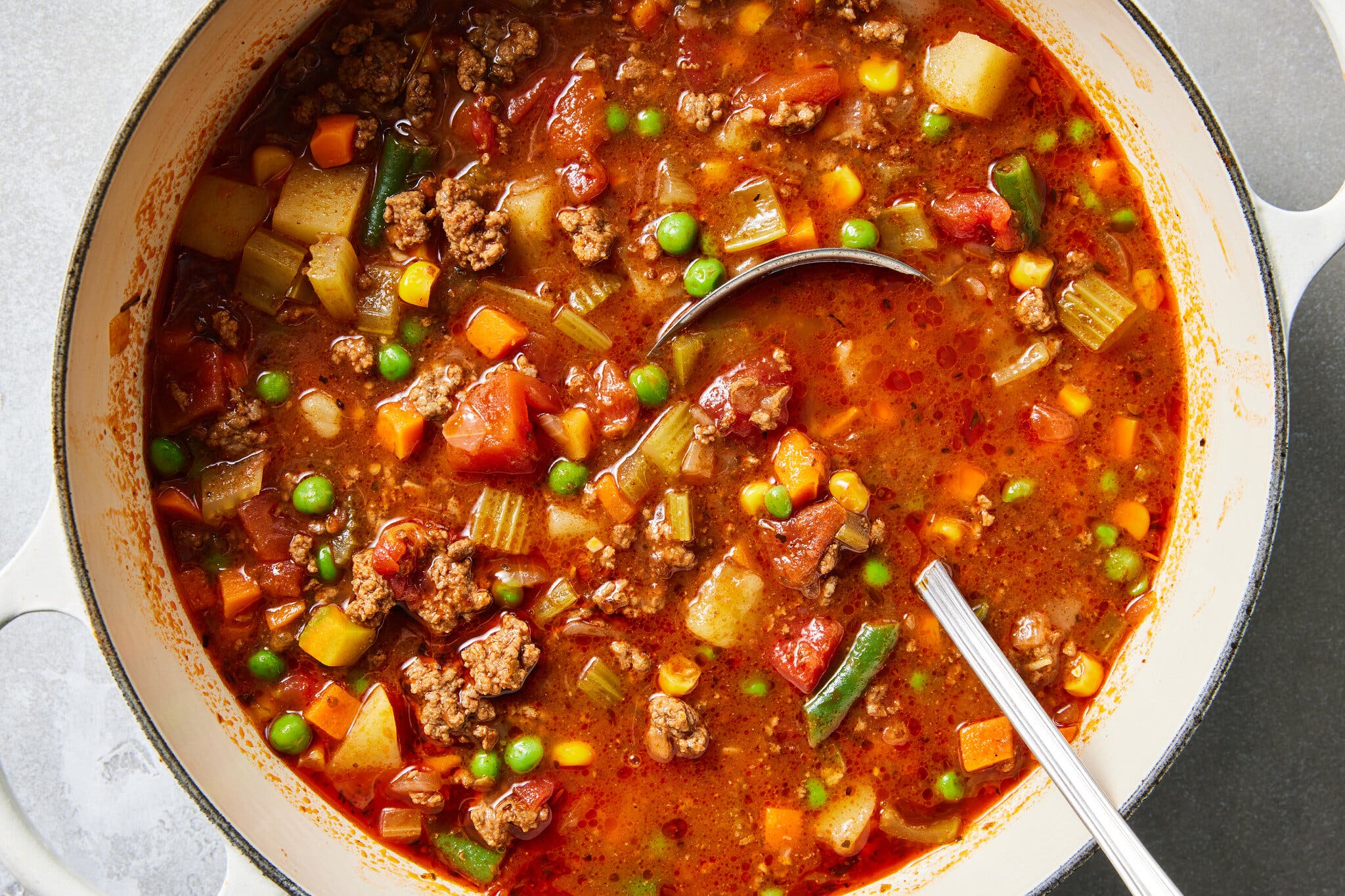 An overhead shot of a white Dutch oven filled with a thick, reddish-brown soup containing chunks of ground beef, peas, corn, carrots, potatoes, and celery; a silver ladle rests inside the pot.