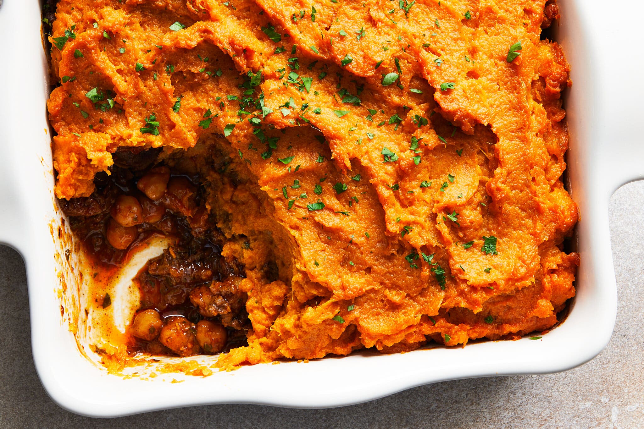 An overhead view of a white baking dish containing a savory shepherd’s pie; the bright orange sweet potato topping, sprinkled with parsley, is scooped away to reveal a brown, saucy ground beef and chickpea filling.