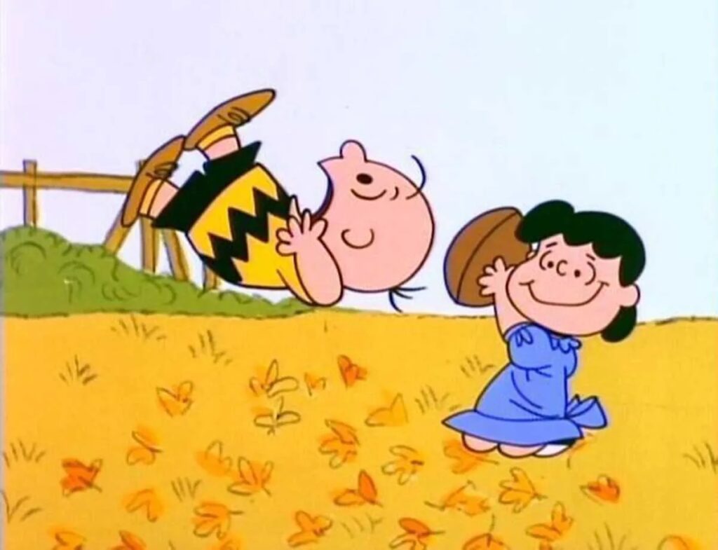 A cartoon boy in a yellow zigzag shirt falls backward in midair as a girl in a blue dress smiles, holding a football. They are on a grassy field with scattered orange leaves.