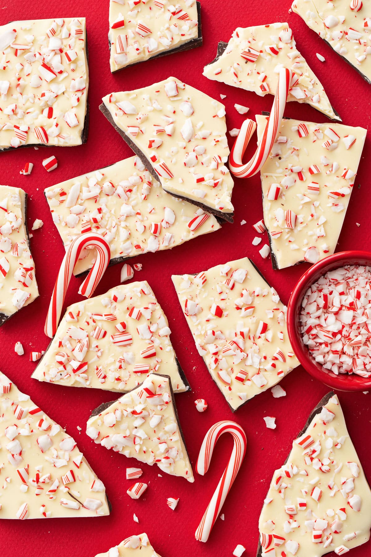 Overhead view of rough broken pieces of homemade peppermint bark scattered on a red background with bits of crushed candy canes scattered around.