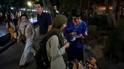 NurPhoto via Getty Images During a nighttime walk in northern Tehran, two young women are standing beside a small display of handmade pottery. One of them is entering information into a customer’s mobile phone. Pedestrians pass by, while the streetlights and surrounding trees illuminate the scene.
