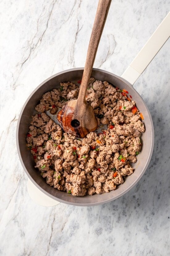 This hearty white bean soup with turkey sausage and kale is packed with protein and fiber, full of cozy flavor, and perfect for chilly days! Browning turkey sausage in skillet with wooden spoon.