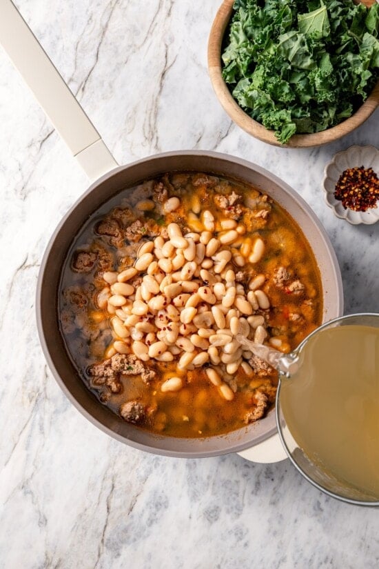 This hearty white bean soup with turkey sausage and kale is packed with protein and fiber, full of cozy flavor, and perfect for chilly days! Pouring chicken stock into pot of white bean soup.